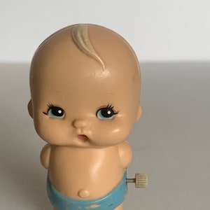Może przedstawiać: Zabawka wijąca się, przedstawiająca dziecko w niebieskim pieluszce i z białym kluczykiem do nakręcania. Dziecko ma jasnobrązową cerę, niebieskie oczy i jasnobrązowy przedziałek.