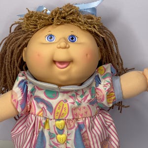May include: A vintage Cabbage Patch Kids doll with brown yarn hair and blue eyes. The doll is dressed in a patterned dress with pink and white stripes, blue shorts, and a blue bow. The dress has yellow heart-shaped buttons.