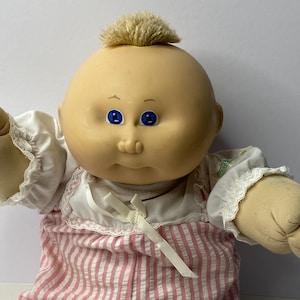 Könnte beinhalten: Eine Cabbage Patch Kid Puppe mit blonden Haaren, blauen Augen und einem rosa-weiß gestreiften Outfit. Die Puppe sitzt mit gekreuzten Beinen und ausgestreckten Armen.