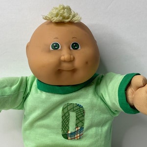 以下が含まれることがあります： ブロンドの髪と緑色の目をしたキャベツパッチキッズのドール。緑色のシャツを着ており、前面にパッチワークの数字「0」が縫い付けられています。