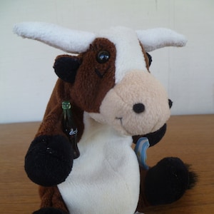 May include: A plush toy cow with brown and white fur, black hooves, and a white belly. The cow is holding a small Coca-Cola bottle. It has a tag attached and is sitting on a wooden surface.