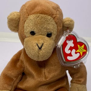 May include: A brown plush monkey toy with a soft, textured body and a heart-shaped tag. The monkey has a light brown face, ears, hands, and feet, with black eyes and a V-shaped nose. The tag reads "Beanie Original Baby".