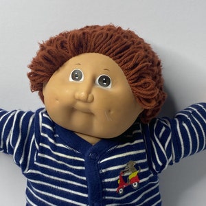 May include: A Cabbage Patch Kid doll with brown yarn hair wearing a blue and white striped sleeper with a red scooter and monkey applique.