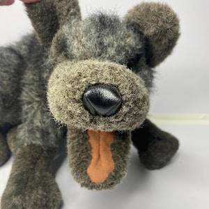 Puede incluir: Un juguete de peluche con forma de perro con pelaje texturizado gris y marrón. El perro tiene una nariz de cuero negro y una lengua naranja. El juguete está tumbado, con las patas delanteras visibles. Las orejas son redondas.