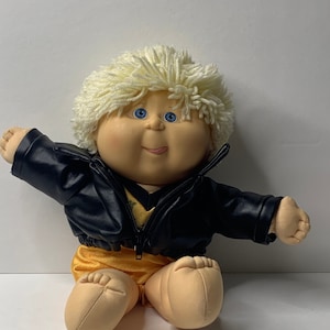 Könnte beinhalten: Eine Cabbage Patch Kid Puppe mit blonden Haaren, blauen Augen und einem breiten Grinsen. Die Puppe trägt eine schwarze Lederjacke und eine gelbe Shorts.