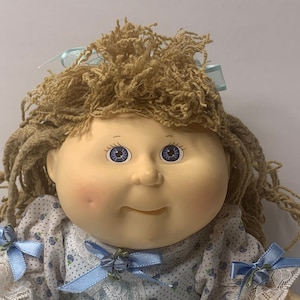 Könnte beinhalten: Eine Cabbage Patch Kid Puppe mit blonden Garnhaaren, blauen Augen und einem hellblauen und weißen Blumenkleid mit blauen Schleifen.