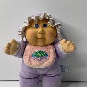 May include: A purple Cabbage Patch Kid doll wearing a pink bib with the text "BabyLand" and a white and purple hat. The doll has blue eyes and a rosy complexion.