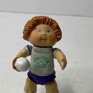 Puede incluir: Una figura de juguete Cabbage Patch Kids que lleva un uniforme deportivo blanco y morado con un logotipo verde que dice "Cabbage Patch Kids". La figura sostiene una pelota blanca en su mano derecha.