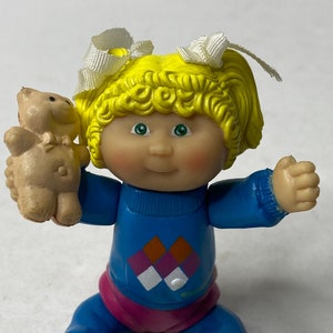 Op de afbeelding: Een plastic Cabbage Patch Kid pop met blond krullend haar, die een blauwe trui met een ruitpatroon, een roze broek en witte schoenen draagt. De pop houdt een kleine bruine teddybeer vast.