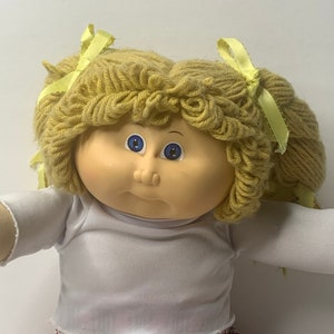 Può includere: Una bambola vintage con capelli biondi di lana a codini, ognuno legato con un nastro giallo. La bambola ha gli occhi blu e un'espressione neutra. Indossa una maglietta bianca a maniche lunghe e una gonna a quadri rosa. Le braccia della bambola sono tese.