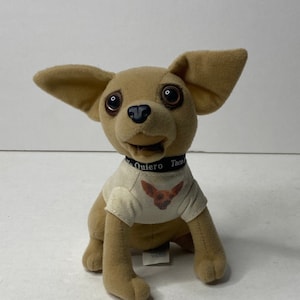 Puede incluir: Un chihuahua de peluche marrón que lleva una camiseta blanca con un gráfico de chihuahua y un collar negro que dice "Yo Quiero Taco Bell".