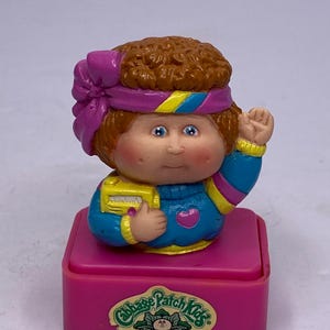 Pode incluir: Uma figura Cabbage Patch Kids com uma base rosa. A boneca tem detalhes em azul e amarelo na faixa de cabeça e nas mangas. A boneca está segurando um objeto amarelo e tem um coração rosa no peito. O logotipo Cabbage Patch Kids está na base.