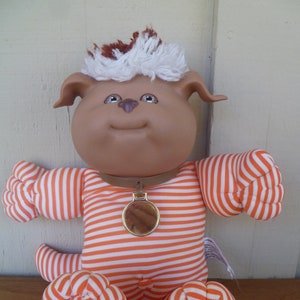 May include: A plush toy dog with brown fur, white fur on its head, and orange and white striped pyjamas. The dog has a brown nose, black eyes, and a gold collar with a tag.