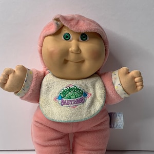 May include: A pink Cabbage Patch Kid doll wearing a pink and white outfit with a bib that says "BabyLand". The doll has green eyes and a rosy face.