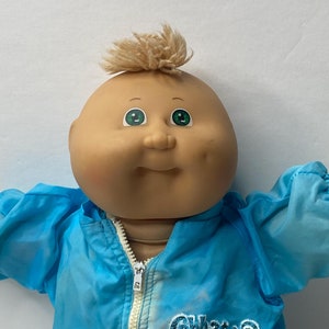 May include: A Cabbage Patch Kid doll wearing a blue jacket with the words "Cabbage Patch Kids" printed on the front. The doll has blonde hair, green eyes, and a rosy complexion.