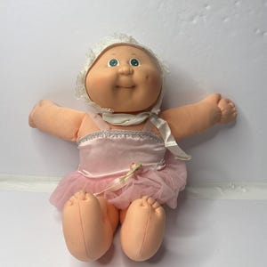 Könnte beinhalten: Eine Vintage-Puppe mit pfirsichfarbenem Gesicht und Gliedmaßen, die ein rosa Ballettkleid mit einem Tüllrock trägt. Die Puppe hat eine weiße Spitzenhaube und ist mit ausgestreckten Armen posiert.