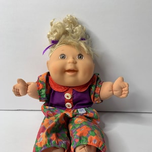 Może przedstawiać: Lalka Cabbage Patch Kid z blondą kręconą czupryną, ubrana w fioletowo-pomarańczowy strój w kwiaty z fioletową kokardką. Lalka ma uśmiechniętą twarz z brązowymi oczami i czerwonymi ustami.