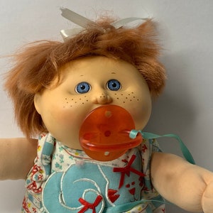 Peut inclure: Une poupée Cabbage Patch Kid aux cheveux roux, aux yeux bleus et aux taches de rousseur portant une robe bleue et blanche avec un nœud rouge. La poupée tient une sucette rouge.