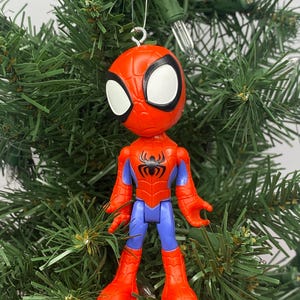 Puede incluir: Un adorno navide&ntilde;o de Spider-Man. El adorno es rojo, azul y negro, con grandes ojos blancos. La figura est&aacute; sujeta a un alambre plateado. El adorno est&aacute; colocado sobre un fondo de ramas verdes de &aacute;rbol de Navidad.