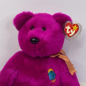 Puede incluir: Un oso de peluche magenta con una nariz negra y ojos negros pequeños. Tiene una cinta dorada y un parche bordado con un diseño de globo y el número 2000. Una etiqueta en forma de corazón está unida a la oreja. Un juguete de peluche.