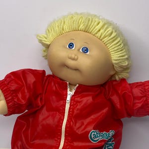 May include: A vintage Cabbage Patch Kids doll with a blonde yarn hairstyle and blue eyes. The doll wears a red nylon jacket with a white zipper and the words "Cabbage Kids" in green. The doll is wearing blue denim pants.