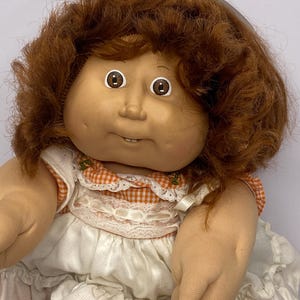 May include: A vintage doll with a soft, tan face, brown eyes, and auburn hair. The doll wears a white dress with orange gingham trim and a white ribbon in its hair. The doll's arms and legs are made of a soft, flesh-toned material.