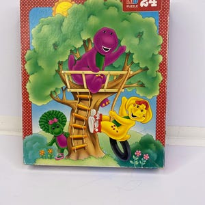 May include: A rectangular puzzle box featuring a purple dinosaur sitting in a treehouse, a yellow dinosaur on a tire swing, and a small green dinosaur standing below. The box has a red border with white dots and the text MB PUZZLE 24 in the corner.