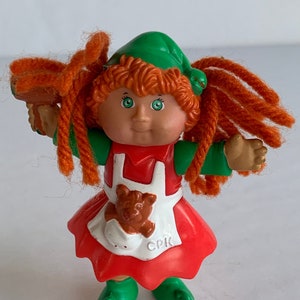 Op de afbeelding: Een rode en groene Cabbage Patch Kid pop die is verkleed als elf. De pop heeft rood garen haar, een groene hoed en een rode jurk met een witte schort. De pop houdt een bruine teddybeer in haar armen. De pop draagt groene schoenen.