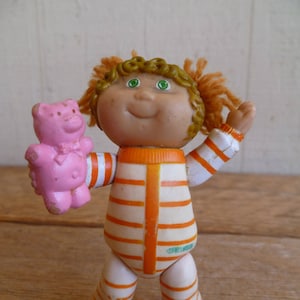 Op de afbeelding: Een Cabbage Patch Kid pop die een oranje en wit gestreept pyjama draagt, houdt een roze teddybeer vast. De pop heeft groene ogen en bruin haar.