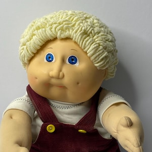 May include: A Cabbage Patch Kid doll with blonde yarn hair wearing a yellow knitted hat and brown corduroy dungarees with yellow buttons.