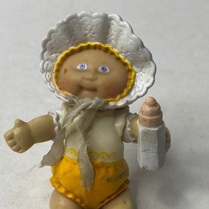 Puede incluir: Una pequeña muñeca de plástico Cabbage Patch Kid que lleva un gorro blanco con ribete amarillo, un vestido amarillo con ribete de encaje blanco y zapatos amarillos. La muñeca sostiene un biberon blanco.