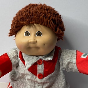May include: A vintage doll with a round face, brown eyes, and curly brown hair. The doll is wearing a silver and red outfit with red buttons and white trim. The doll's arms are outstretched.