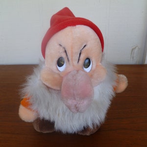 May include: A Grumpy soft toy from Snow White. The doll has a red hat, a large pink nose, and a white beard. His face has an angry expression. The doll's body is a light peach colour, with brown shoes and orange sleeves.