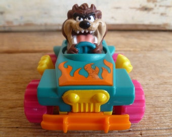 May include: A teal, orange, and yellow toy car with a cartoon Tasmanian Devil figure behind the wheel. The car has flames painted on the hood and a grill with two yellow headlights.