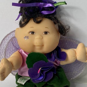 May include: A Cabbage Patch Kids doll dressed as a fairy with purple wings, a purple flower crown, and a green and purple flower dress. The doll has brown eyes, black hair, and a painted flower on its cheek. The doll is sitting on a white surface.