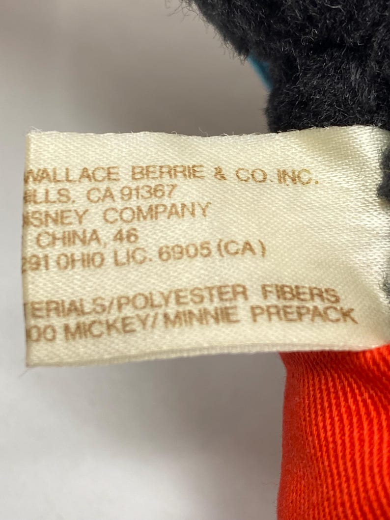 May include: A white tag with black text that reads "WALLACE BERRIE & CO. INC. ELLS, CA 91367 SNEY COMPANY CHINA, 46 291 OHIO LIC. 6905 (CA) ERIALS/POLYESTER FIBERS 00 MICKEY/MINNIE PREPACK"