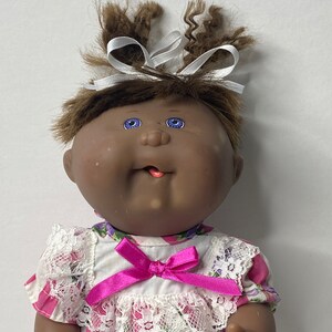 Könnte beinhalten: Eine Cabbage Patch Kid Puppe mit braunen Haaren, blauen Augen und einem rosa-weiß gestreiften Kleid mit einem weißen Spitzenkragen und einer rosa Schleife. Die Puppe hat eine rosa Zunge herausgestreckt.
