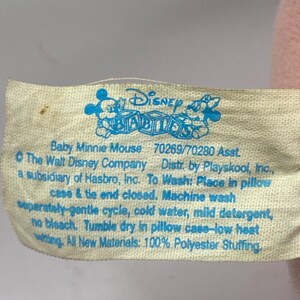 Peut inclure: Gros plan sur une &eacute;tiquette cr&egrave;me d'un jouet en peluche Disney Babies. L'&eacute;tiquette pr&eacute;sente du texte et des graphiques bleus, dont le logo Disney Babies et des instructions de lavage. Le texte indique "Baby Minnie Mouse".
