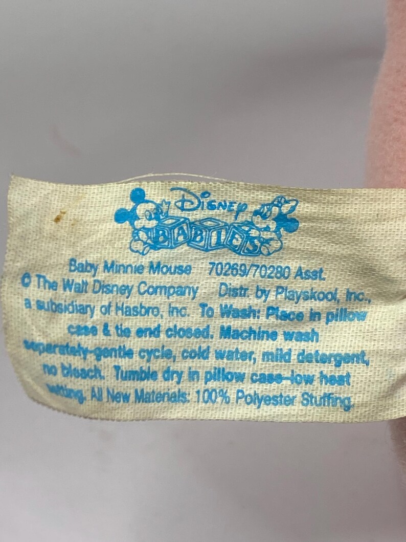 Peut inclure: Gros plan sur une &eacute;tiquette cr&egrave;me d'un jouet en peluche Disney Babies. L'&eacute;tiquette pr&eacute;sente du texte et des graphiques bleus, dont le logo Disney Babies et des instructions de lavage. Le texte indique "Baby Minnie Mouse".