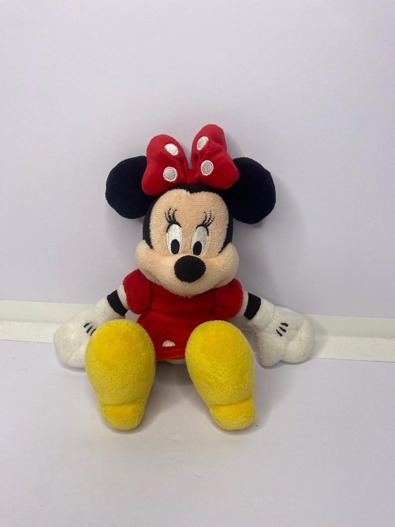 Pu&ograve; includere: Una bambola di peluche di Minnie Mouse, con un vestito rosso, scarpe gialle e un fiocco rosso a pois bianchi. La bambola ha orecchie nere, un naso nero e guanti bianchi. Il giocattolo &egrave; per bambini.