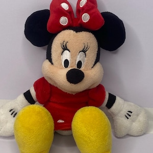Pu&ograve; includere: Una bambola di peluche di Minnie Mouse con un vestito e un fiocco rossi. La bambola ha orecchie nere, naso nero e guanti bianchi. Sono visibili i piedi gialli della bambola.