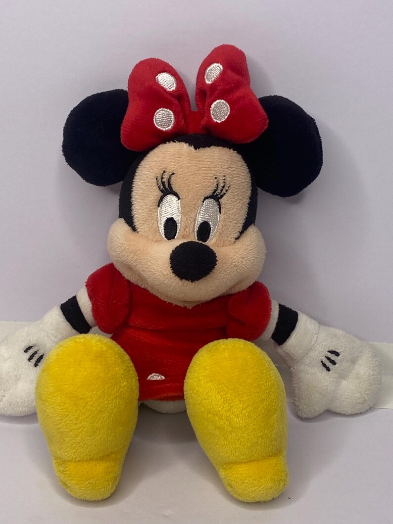 Pu&ograve; includere: Una bambola di peluche di Minnie Mouse con un vestito e un fiocco rossi. La bambola ha orecchie nere, naso nero e guanti bianchi. Sono visibili i piedi gialli della bambola.