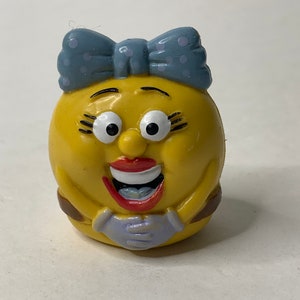 May include: A yellow plastic toy with a blue bow, brown belt, and a smiling face. The toy has large eyes, red lips, and white teeth.
