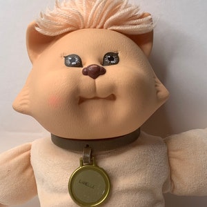 Peut inclure: Un jouet en peluche en forme de chat avec un collier brun clair et une étiquette dorée sur laquelle est inscrit "LANELLE". Le chat a un visage rose clair avec des yeux bruns et un nez brun clair. Le chat a une fourrure brun clair et blanche.