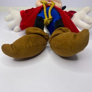 May include: A plush toy of a cartoon character, featuring a cream-colored face, black ears, and a black topknot. The character wears a red cape, blue shorts, and brown shoes. White gloves are visible. A gold-colored rope is around the neck.