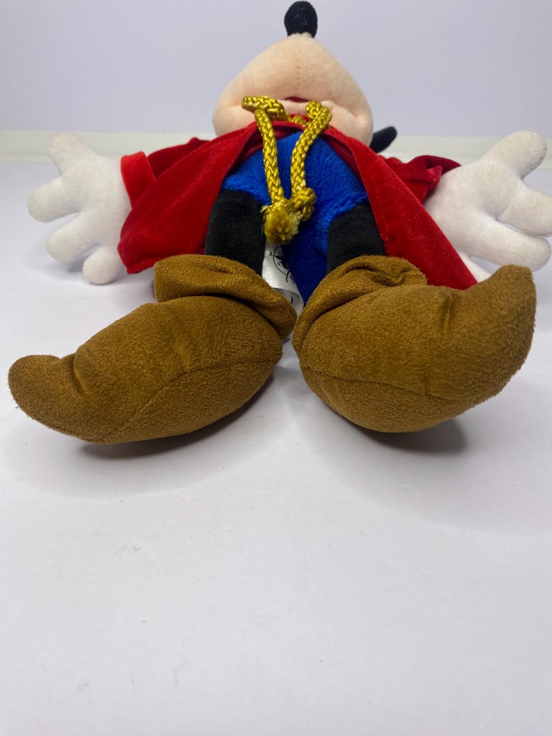 May include: A plush toy of a cartoon character, featuring a cream-colored face, black ears, and a black topknot. The character wears a red cape, blue shorts, and brown shoes. White gloves are visible. A gold-colored rope is around the neck.