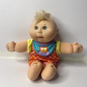Könnte beinhalten: Eine Cabbage Patch Kid Puppe mit blonden Haaren, grünen Augen und einem rosa-orangenen karierten Outfit. Die Puppe trägt ein blaues Lätzchen mit einem Eulenmotiv und dem Wort "smile".