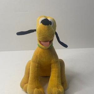 May include: A yellow plush toy dog with black ears and a green collar. The dog is sitting with its tongue sticking out.