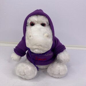 May include: A plush white hippo wearing a purple hooded sweatshirt. The sweatshirt has the word "LUMS" and the word "Lulu" embroidered on the front. The hippo has red eyes and is sitting upright.