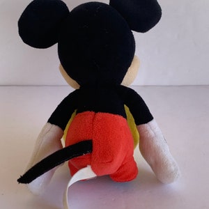 Op de afbeelding: Een pluche Mickey Mouse-speelgoed, gezien van achteren, met zwarte oren, hoofd en staart. Het speelgoed heeft een rode onderkant, gele accenten en witte handschoenen. Een wit label is aan het speelgoed bevestigd.